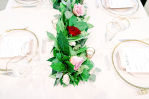 Wedding Reception Feasting Table with White Linen Tablecloth and Clear Glass Charger Plates with Gold Rim and Gold Rimmed Glasses | White Blush Pink and Maroon Red Roses Centerpiece Runner with Eucalyptus Greenery