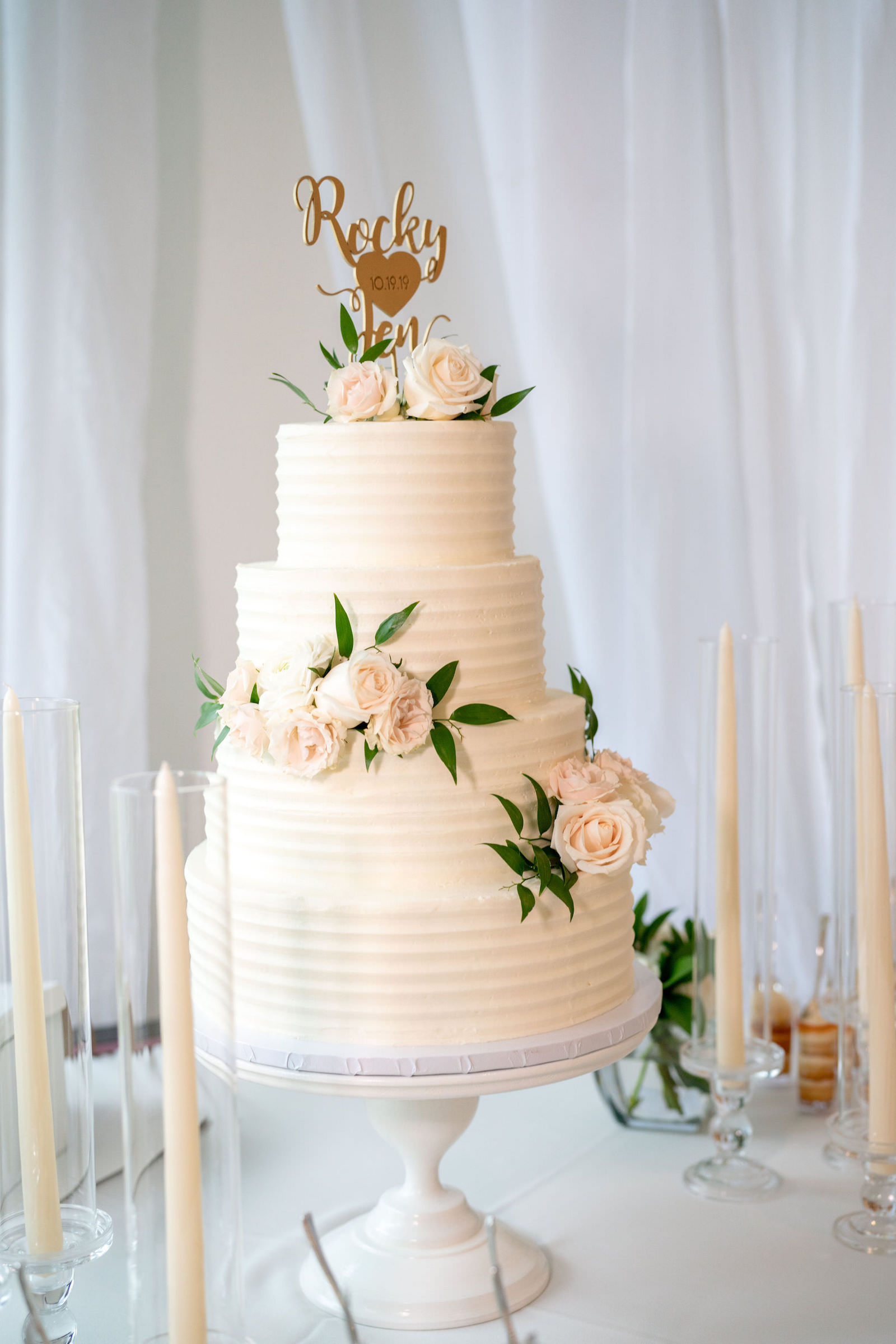 Four Tier Wedding Cake with Ribbed Textured Buttercream Icing and Fresh Flowers Accents of Roses and Greenery with Gold Die Cut Names Cake Topper | Wedding Cake Table with White Round Pedestal Cake Stand and Taper Candles