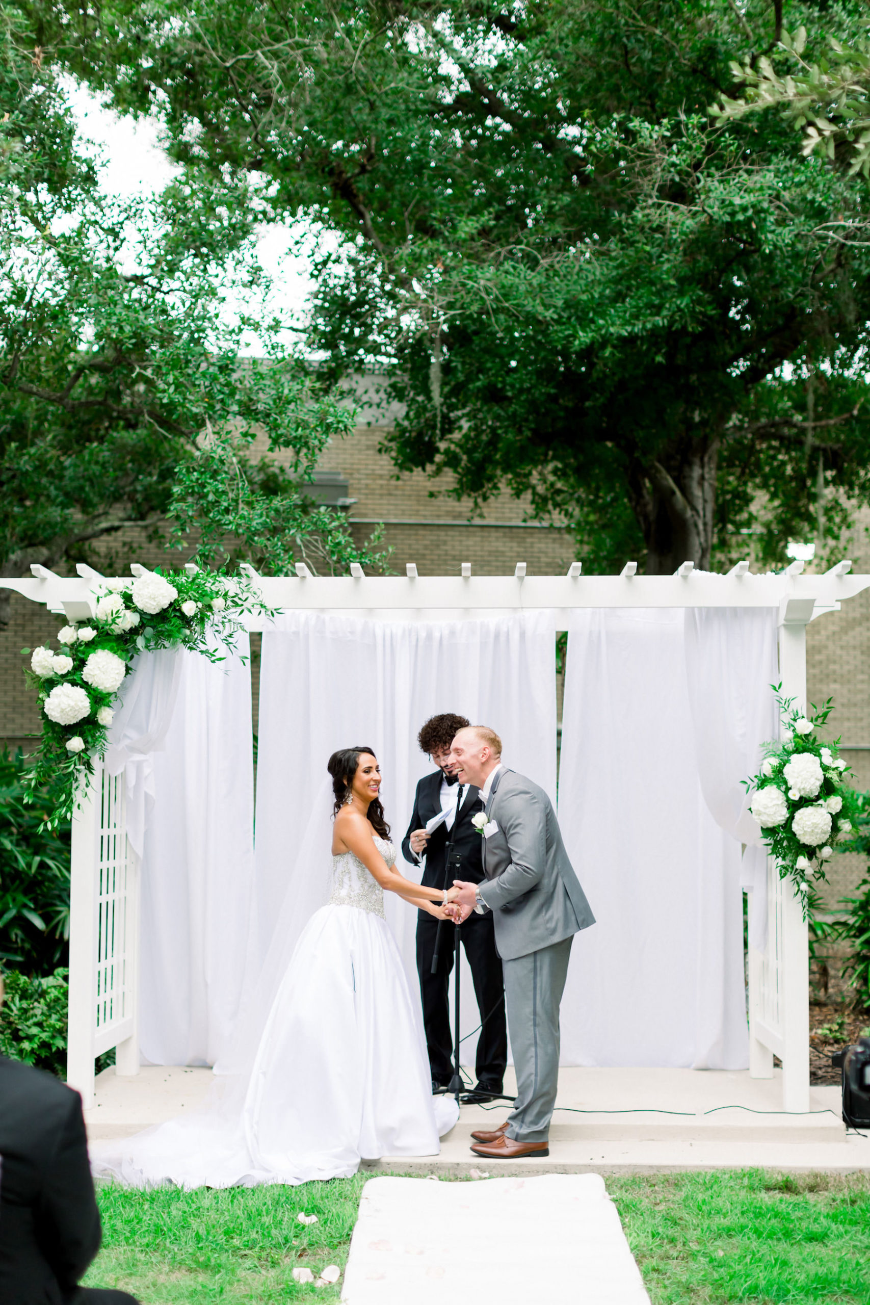 Romantic Bride and Groom Exchanging Vows Wedding Ceremony Portrait Under White Arbor Arch with Hydrangeas and Greenery Floral Arrangements and White Draping | Wedding Venue Tampa Garden Club | Wedding Photographer Shauna and Jordon Photography | Wedding Dress Truly Forever Bridal