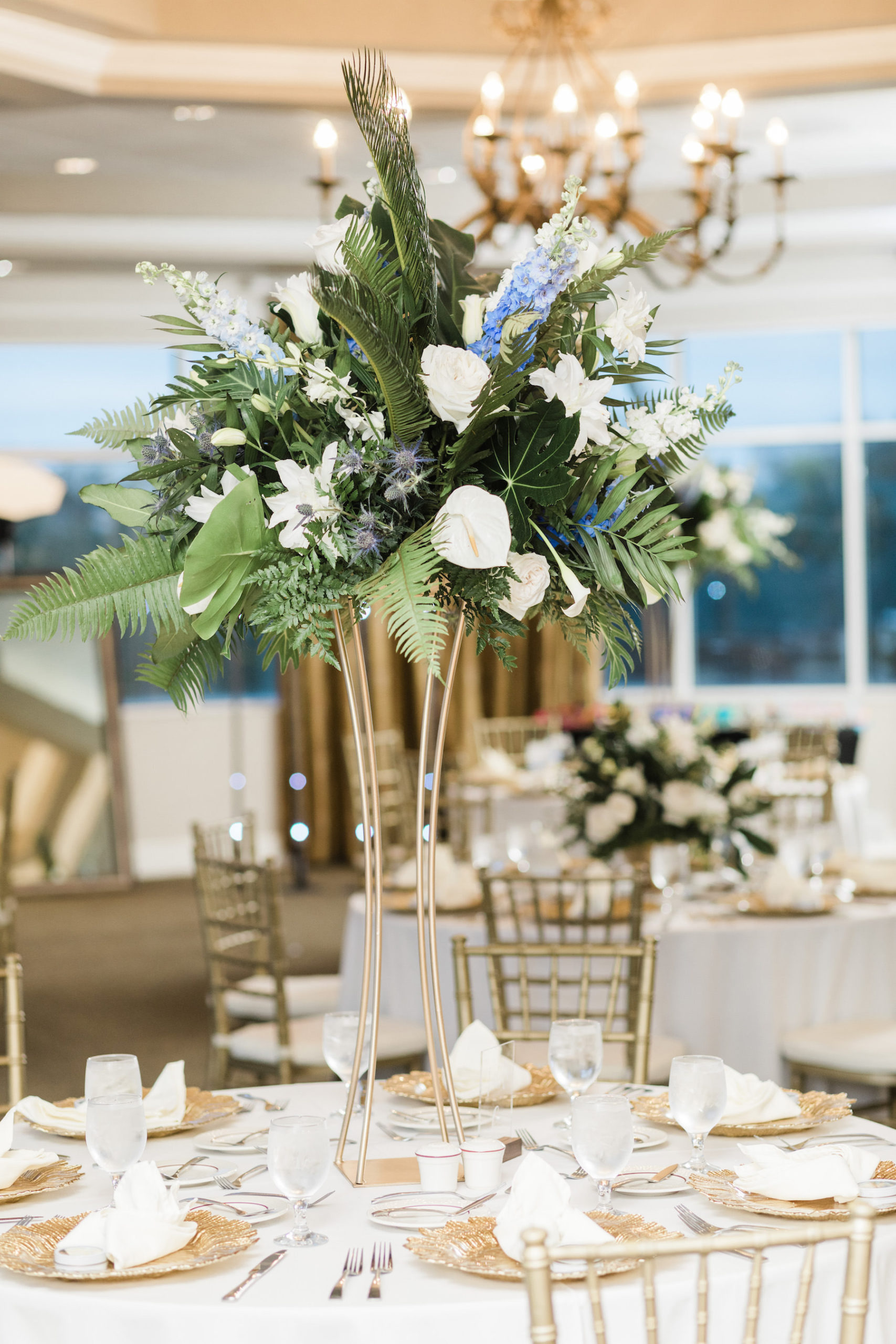 Indoor Ballroom Reception with Gold Chargers, Chiavari Chairs and Tall Tropical Centerpieces with Greenery, White and Blue Flowers
