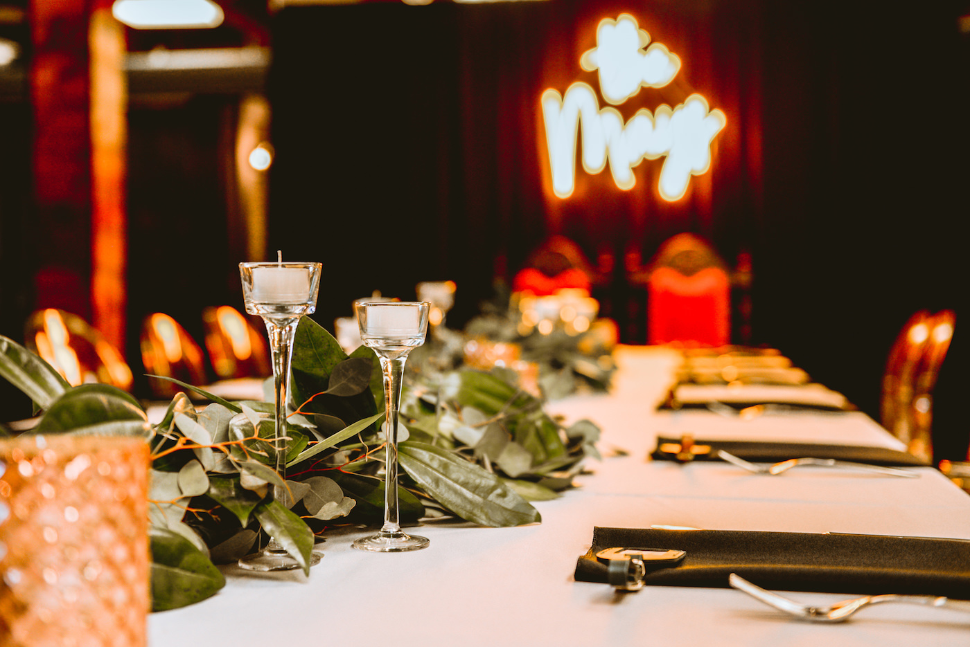 Wedding Reception Feasting Table with Clear Ghost Chairs and Greenery Garland Centerpiece with Tea Light Votive Candles and Black Napkins | Bride and Groom Vintage Chairs with Neon Sign Backdrop | St. Pete Wedding Venue Station House