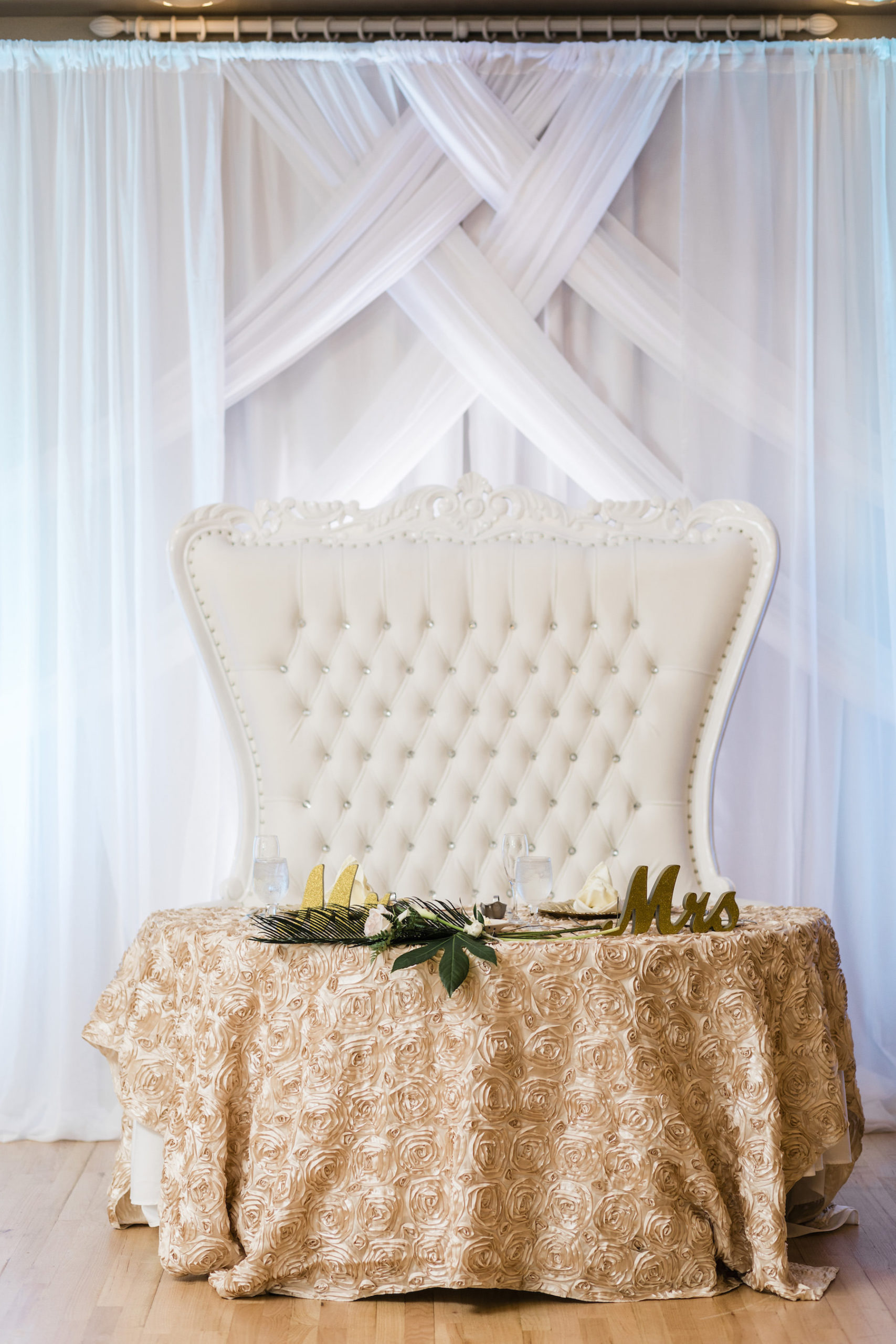 Wedding Sweetheart Table with Ornate Vintage Tufted Love Seat and Champagne Embroidered Textured Rosette Table Linen | White Pipe and Drape Backdrop with Uplighting | Mr and Mrs Sweetheart Table Sign