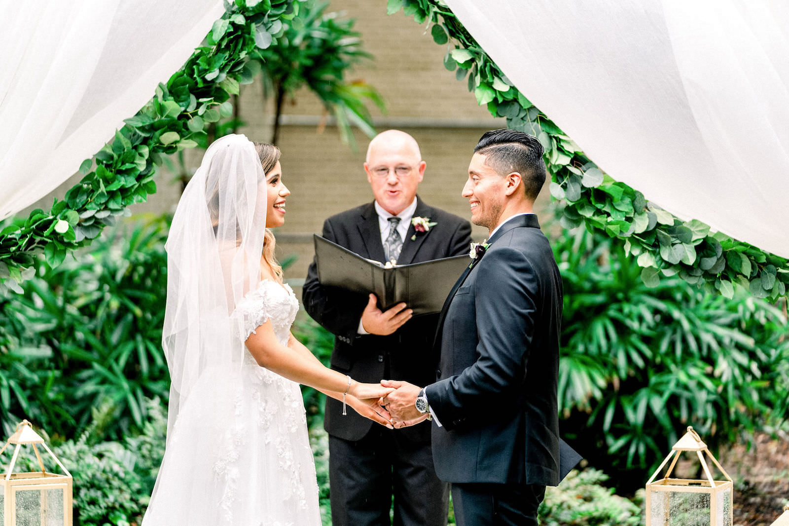 Bride and Groom Exchanging Vows during Garden Wedding Ceremony with Draped Arbor and Greenery Backdrop