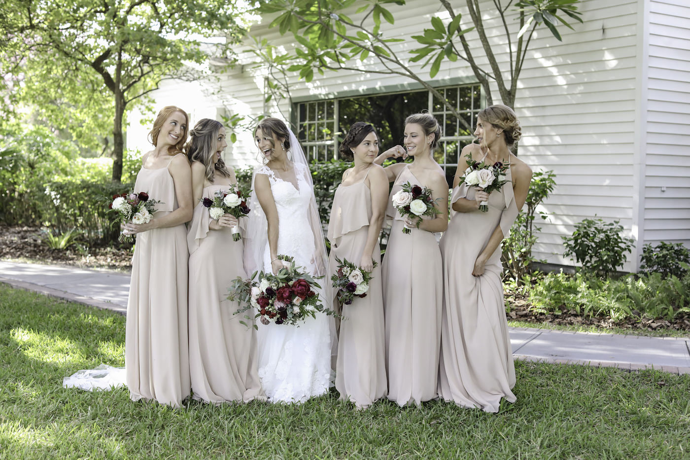 Bride in Lace Wedding Dress with Bridesmaids in Neutral Nude Dresses Holding Burgundy, Blush Pink and Ivory Roses with Greenery Floral Bouquet | Wedding Photographer Lifelong Photography Studio | Tampa Bay Wedding Planner Blue Skies Weddings and Events | Dress Shop Bella Bridesmaids