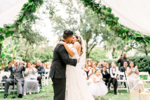 Tampa Garden Club Tampa Wedding Venue | Bride and Groom First Kiss during Garden Wedding Ceremony with Draped Arbor and Greenery Backdrop