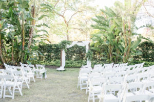 Tampa Wedding Venue the Tampa Garden Club | Outdoor Garden Wedding Ceremony with White Garden Chairs and Wood Arch with White Draping and Greenery Floral Arrangements