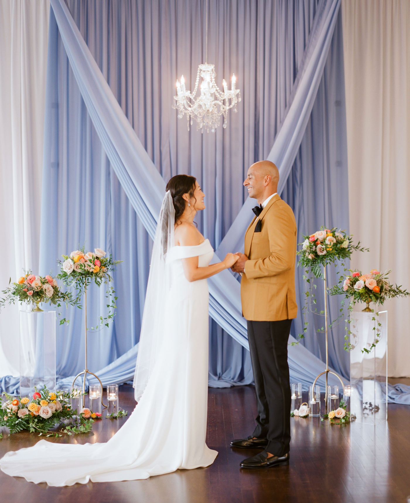 Sophisticated Tampa Bay Bride and Groom Exchange Vows During Intimate Styled Shoot Wedding, Elegant Ceremony Decor with Lush Velvet Blue Draping, Romantic Crystal Chandelier, Decorated with Springtime Inspired Florals, Pink Roses, Peach Carnations, Yellow Flowers, Pops of Gold Accents with Greenery, Bride in Off the Shoulder Long Sheath Wedding Dress, Groom Wearing Mustard Yellow Suit Jacket from The Black Tux | Tampa Bay Wedding Planner Kelly Kennedy Weddings and Events | Florida Luxury Rental Company Kate Ryan Event Rentals