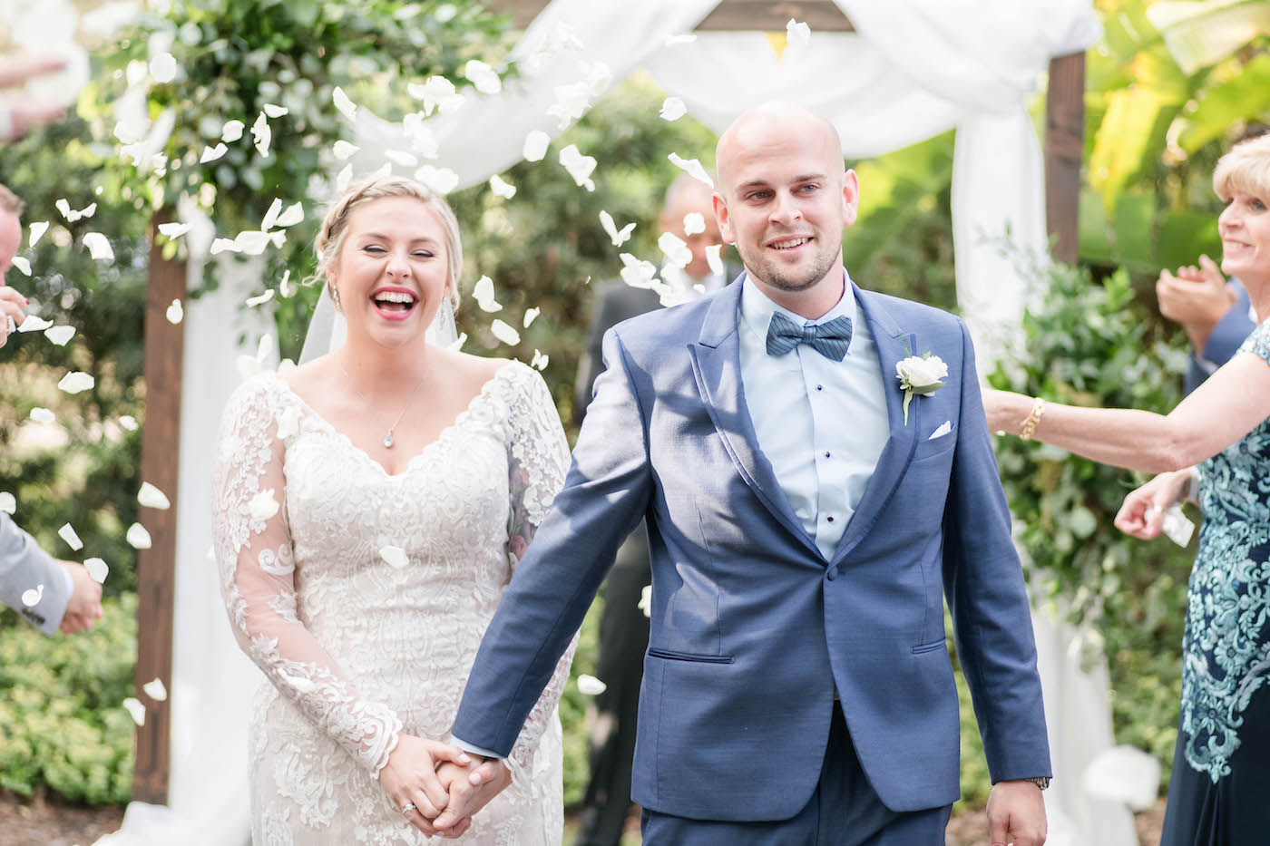 Bride and Groom Walking Down the Aisle in Rose Petal Toss | Ivory Lace Long Sleeve V Neck Bridal Gown and Groom in Blue Suit with Bow Tie