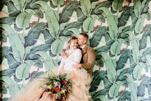 Bohemian Inspired Tampa Bay Bride and Groom Wedding Portrait, In front of Green Palm Leaf Wallpaper, Boho Bride Wearing Oversized Blush Orange Long Tulle Skits, White Lace Long Sleeve Top, Holding Vintage Wildflower Bouquet, with Orange, Purple, Yellow, Red, Eggplant and Ivory Floral Stems, Thistle, Roses | Tampa Bay Wedding Planner Blue Skies Weddings and Events | Downtown St. Petersburg Wedding Photographer Lifelong Photography Studio | Unique Florida Wedding Venue Station House in DTSP