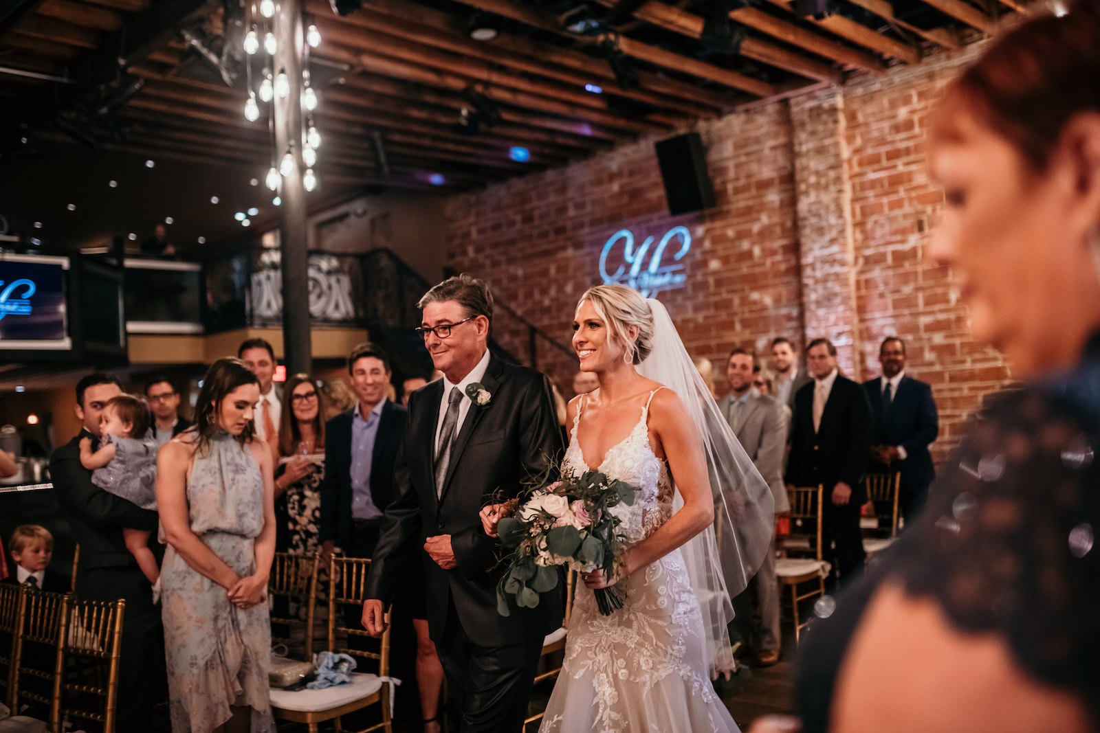 Florida Bride and Father Walk Down the Aisle During Indoor Wedding Ceremony, Interior Summer Ceremony with Exposed Red Brick Wall and String Lighting | Historic Tampa Bay Wedding Venue NOVA 535 in Downtown St. Petersburg