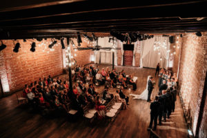 Florida Bride and Groom Exchange Vows During Wedding Ceremony, Interior Summer Ceremony with Exposed Red Brick Wall, Hardwood Floors and String Lighting | Historic Tampa Bay Wedding Venue NOVA 535 in Downtown St. Petersburg