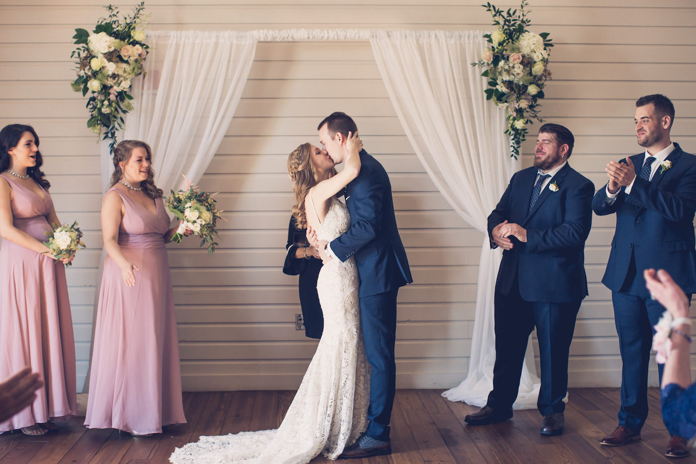 Bride and Groom First Kiss at Tampa Historic Venue with Pipe and Drape Backdrop and White and Blush Pink Rose and Greenery Floral Arrangements | Dusty Rose Bridesmaid Dresses and Navy Blue Groomsmen Suits | Tampa Wedding Photographer Luxe Light Images