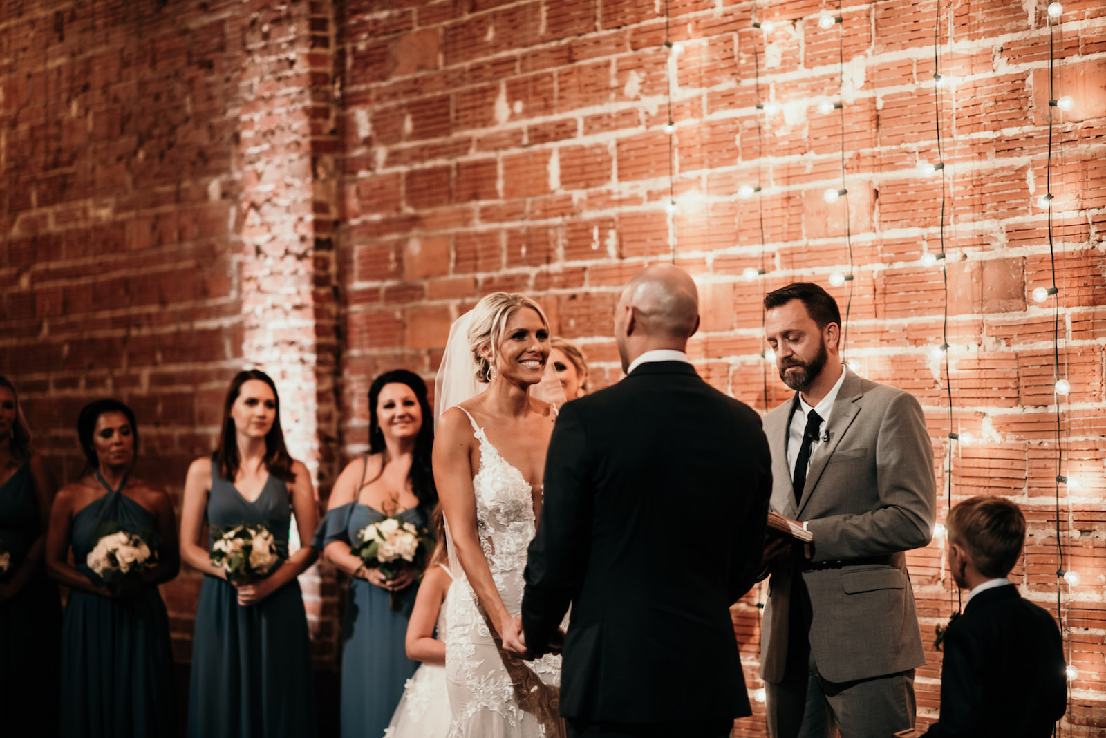 Florida Bride and Groom Exchange Vows During Wedding Ceremony, Interior Summer Ceremony with Exposed Red Brick Wall and String Lighting | Historic Tampa Bay Industrial Wedding Venue NOVA 535 in Downtown St. Petersburg