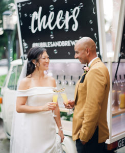 Florida Bride and Groom Cheers at Mobile Champagne and Craft Beer Bar, Bride in Sophisticated Off The Shoulder Gown, Groom Wearing Elegant Mustard Suit Jacket, In Front of Bubbles and Brew | Tampa Bay Wedding Planner Kelly Kennedy Weddings and Events