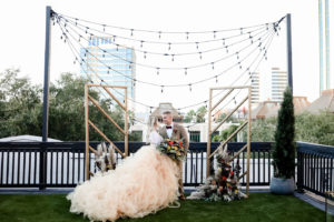 Bohemian Inspired Tampa Bay Bride and Groom Wedding Portrait, In front of Ceremony Backdrop with String Lighting and Outdoor City View, Vintage Wedding Decor with Wooden Alters, Boho Bride Wearing Oversized Blush Orange Long Tulle Skirts, White Lace Long Sleeve Top, Holding Vintage Wildflower Bouquet, with Orange, Purple, Yellow, Red, Eggplant and Ivory Floral Stems, Thistle, Roses, Groom in Brown Suit with Velvet Bowtie | Tampa Bay Wedding Planner Blue Skies Weddings and Events | Downtown St. Petersburg Wedding Photographer Lifelong Photography Studio | Unique Florida Wedding Venue Station House in DTSP