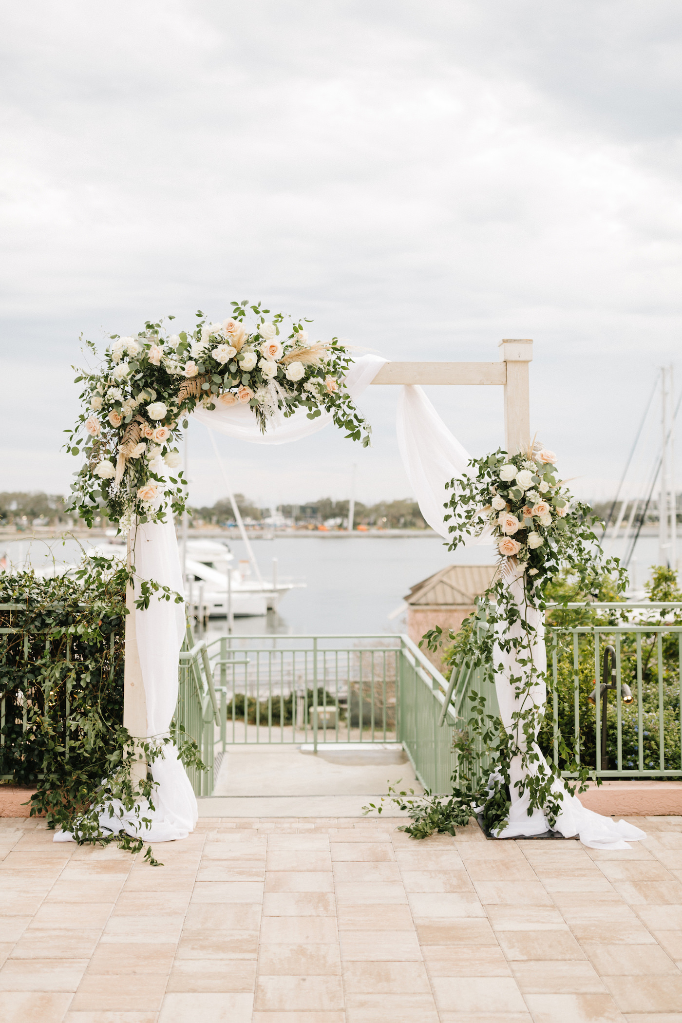 Boho Chic Outdoor Waterfront Ceremony, White Wooden Arch with White Linen Draping, Decorated with Lush Ivory Flowers and Blush Pink Roses, Pampas Grass Bouquets with Greenery | The Esplanade Location of the Vinoy Renaissance Resort in Downtown St. Petersburg | Tampa Bay Luxury Wedding Planner Parties A’ La Carte | Rentals A Chair Affair | Rentals Over the Top Linens