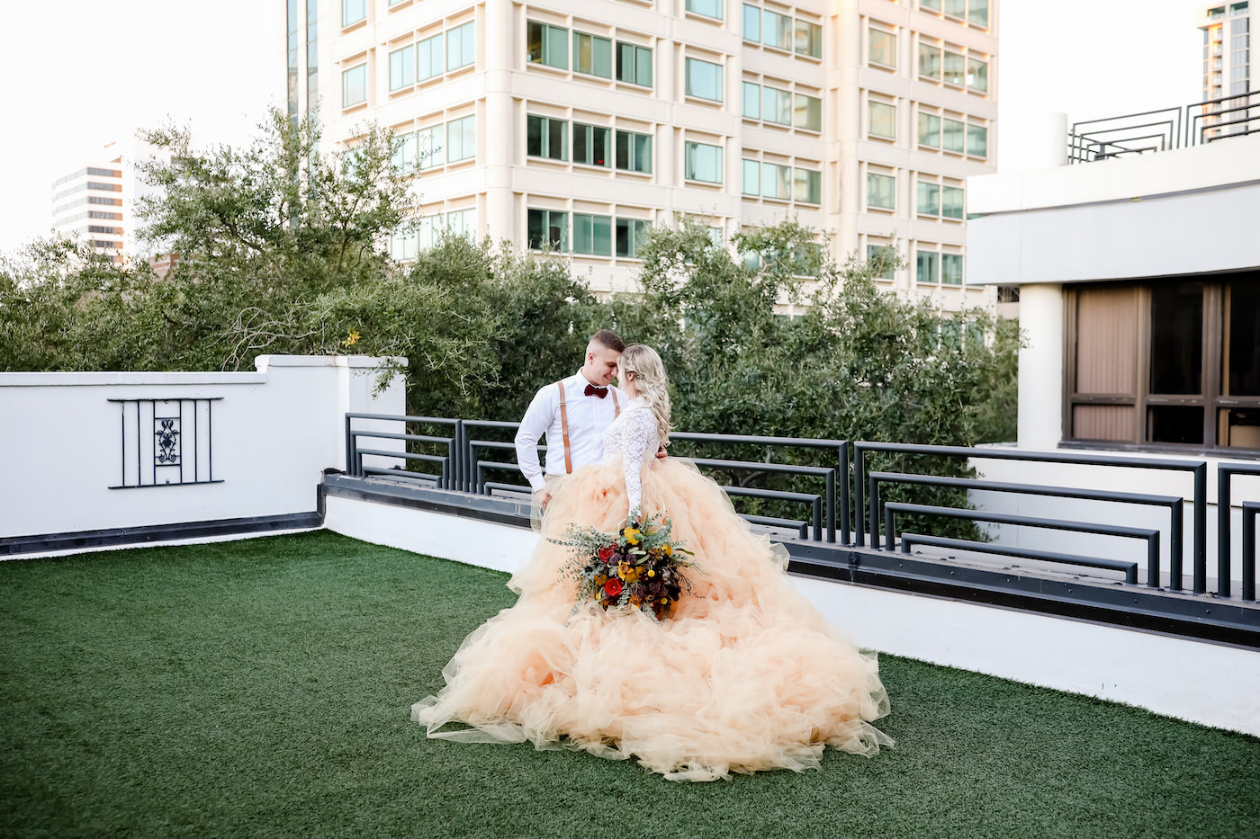 Bohemian Inspired Tampa Bay Bride and Groom Wedding Portrait, In front of Ceremony Backdrop on Outdoor Balcony with City View, Boho Bride Wearing Oversized Blush Orange Long Tulle Skirt, White Lace Long Sleeve Top, Holding Vintage Wildflower Bouquet, with Orange, Purple, Yellow, Red, Eggplant and Ivory Floral Stems, Thistle, Roses, Groom in Brown Suit with Velvet Bowtie | Tampa Bay Wedding Planner Blue Skies Weddings and Events | Downtown St. Petersburg Wedding Photographer Lifelong Photography Studio | Unique Florida Wedding Venue Station House in DTSP