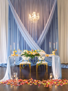 Romantic, Luxurious Wedding Reception and Decor, Sweetheart Table with Light Blue Linens, Acrylic Ghost Table, Gold Metal Chairs. Velvet Draping, Vibrant Pink and Yellow Petals, Peach, Orange, Pink and Red Florals with Greenery, Tall Taper Candles | Florida Wedding Planner Kelly Kennedy Weddings and Event | Kate Ryan Event Rentals