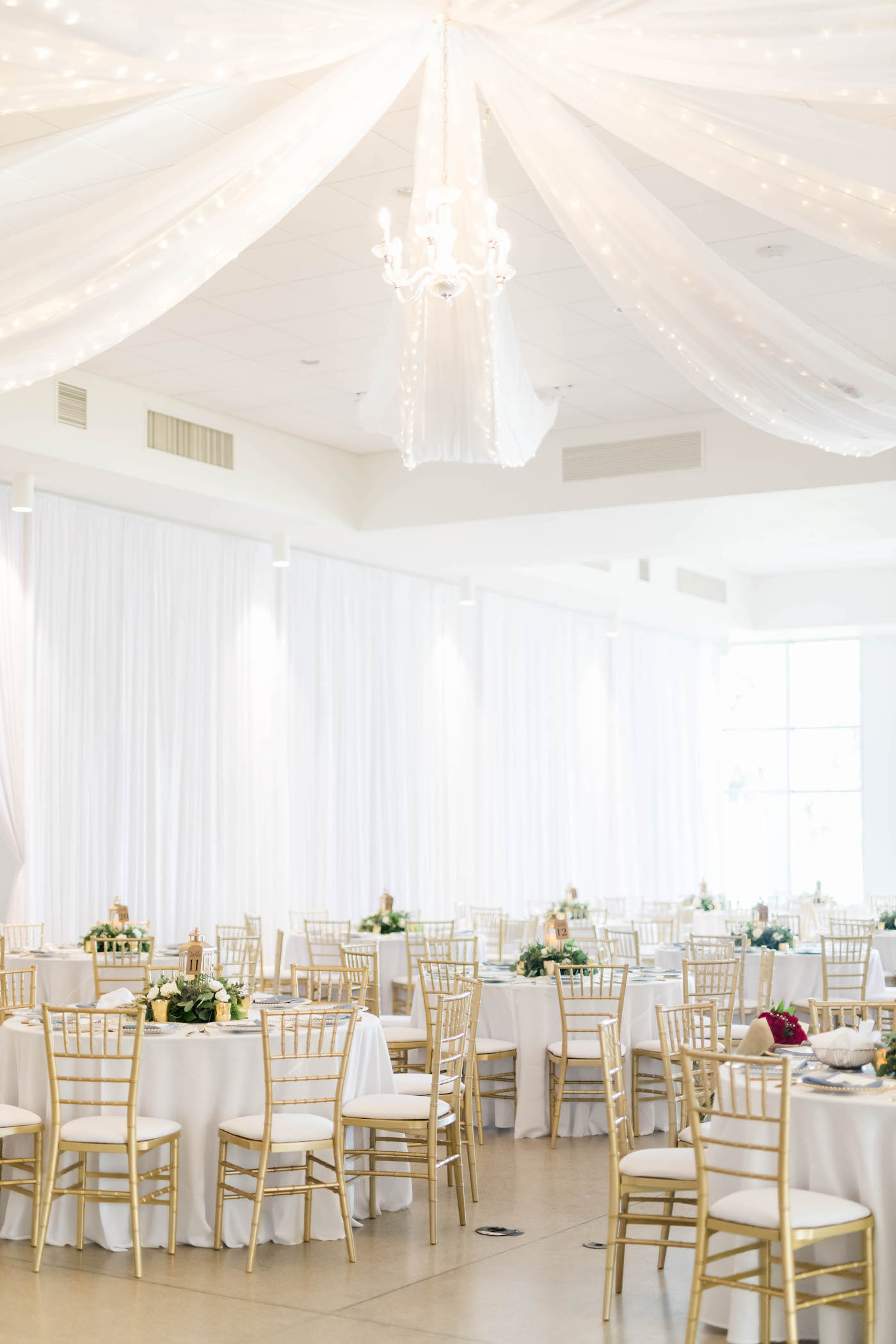 Tampa Wedding Venue the Tampa Garden Club Indoor Reception with Pipe and Drape and Ceiling Draping and Twinkle Lights | Wedding Reception Tables with White Linens and Gold Chiavari Chairs and Low Greenery Centerpieces | Gabro Event Services