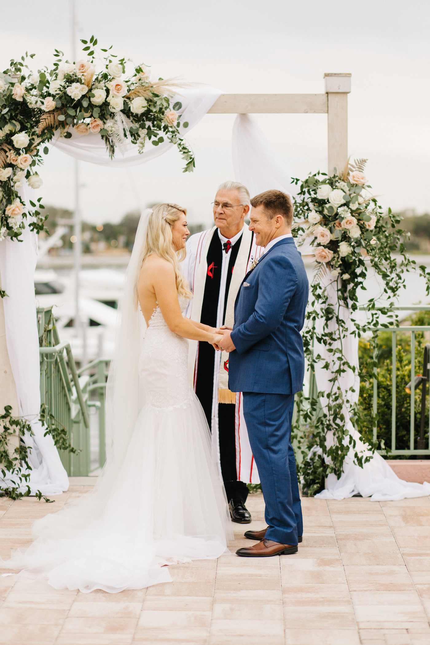 Boho Chic Inspired Wedding Ceremony, Florida Bride and Groom Hold Hands in Outdoor Waterfront Vow Exchange, Ashley Stegbauer Morrison Wearing Modern Ines Di Santo Wedding Dress, White Lace and Tulle Fit and Flare with Plunging Neckline, Altar Decorated in Boho Chic Inspired Ivory Flowers and Blush Pink Roses, Pampas Grass with Greenery, White Wooden Arch with Draping | Downtown St. Pete Wedding Planner Parties A’ La Carte | Luxury Florida Wedding and Bridal Dress Shop Isabel O’Neil Bridal Collection | Wedding Venue The Esplanade Location of the Vinoy Renaissance Resort in Downtown St. Petersburg | Rentals Over the Top Linen Rentals