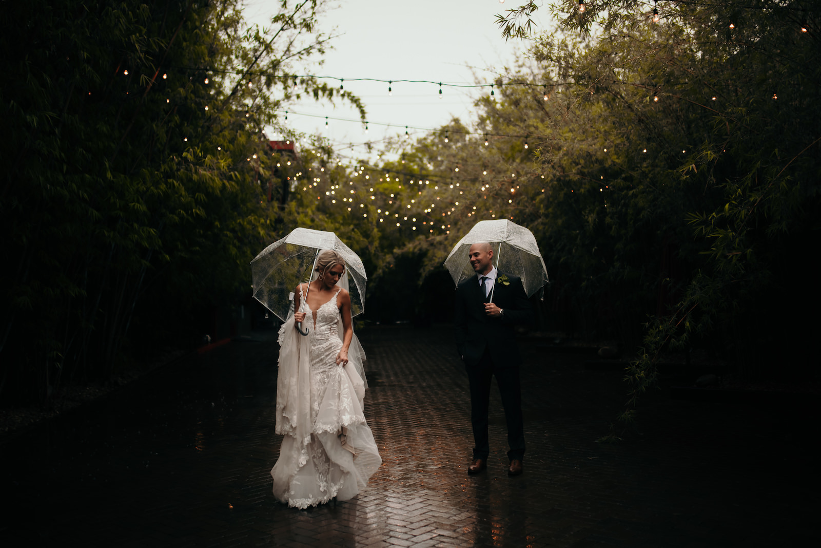 Editorial Inspired Wedding Portrait, Tampa Bay Bride and Groom in Enchanted Brick Paved Courtyard with String-lighting and Bamboo, While Raining, Holding Clear Umbrellas | Unique Florida Wedding Venue NOVA 535