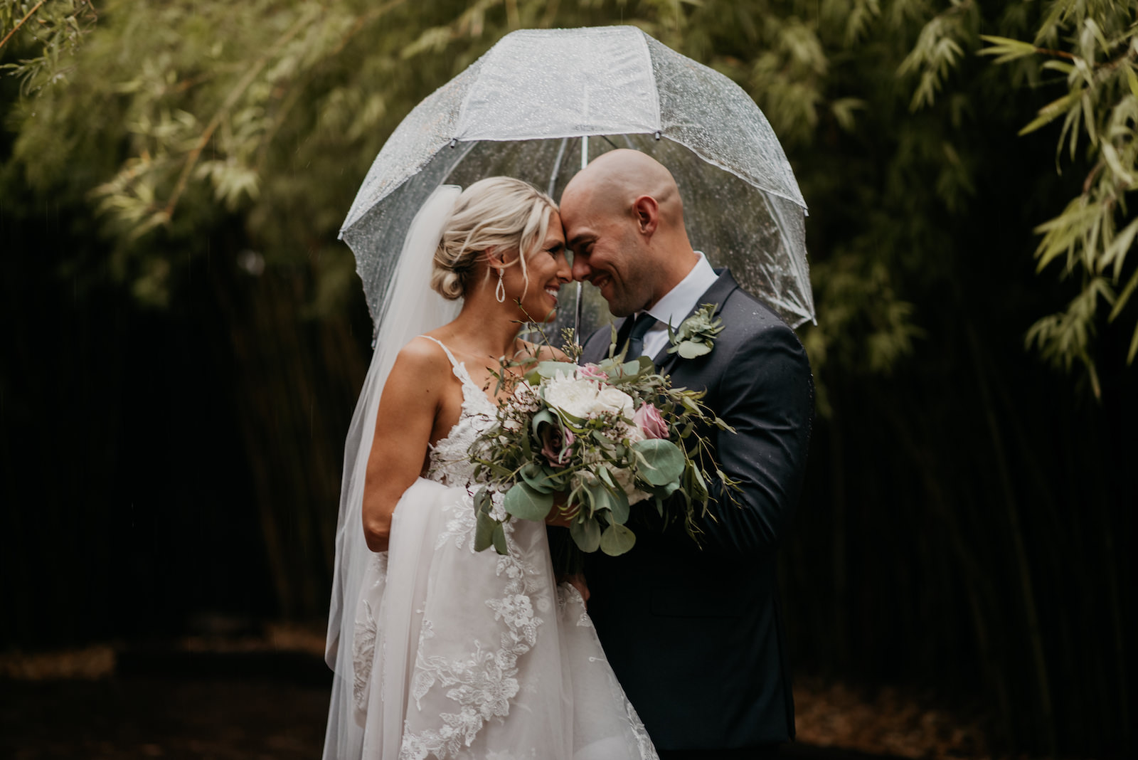 Downtown St. Pete Bride and Groom Wedding Portrait in The Rain, Holding Romantic Bridal Bouquet with White and Pink Roses, and Greenery Eucalyptus, Under Clear Umbrella | Historic Florida Wedding Venue NOVA 535