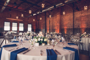 Tampa Wedding Reception Tables with Cobalt Blue Napkins and Greenery Floating Candle Centerpieces in Historic Venue Armature Works Unique Architecture with Brick Walls and Exposed Beam ceilings with Edison Bulb Chandeliers