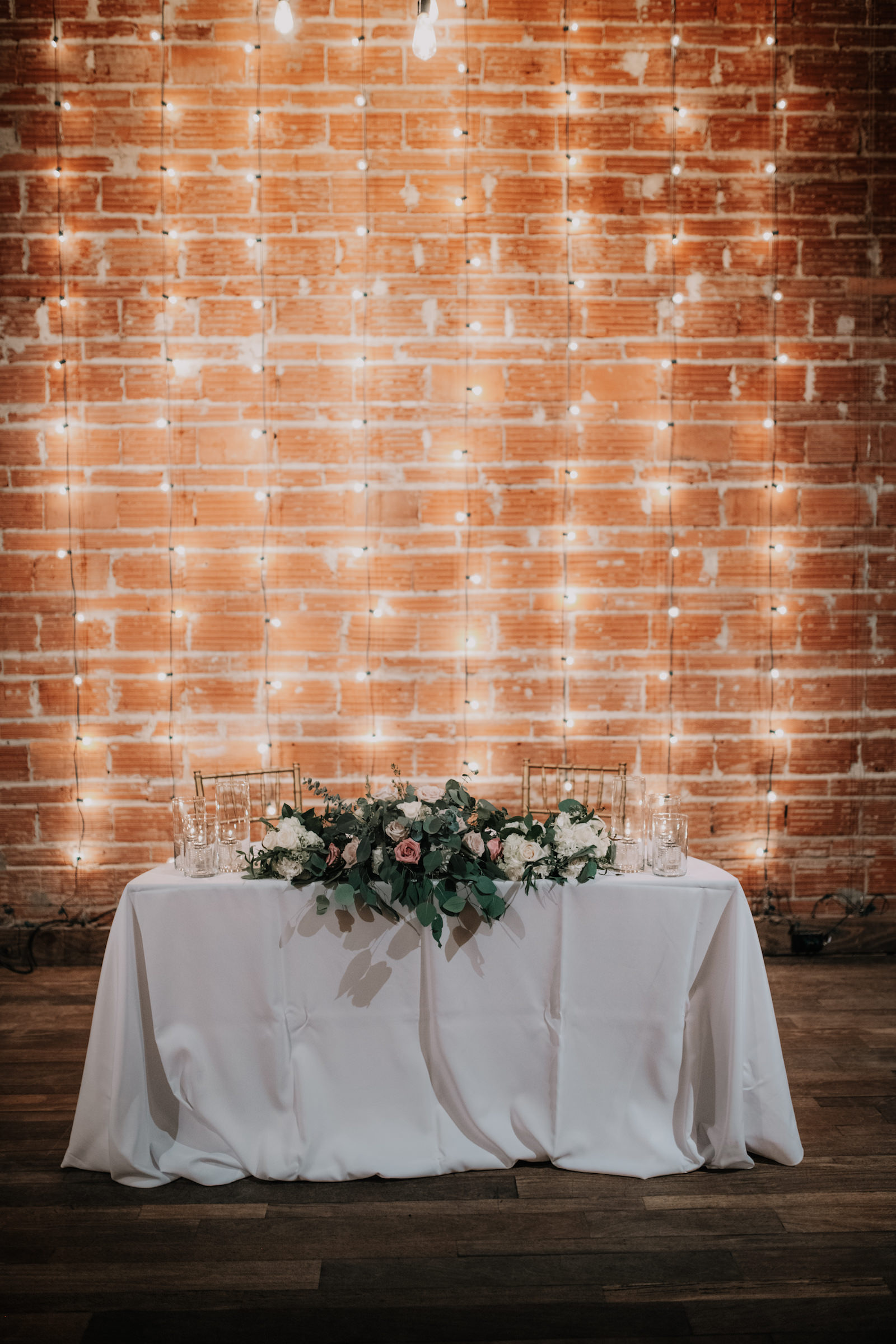 Elegant Wedding Reception Sweetheart Table, White Linens with Gold Chiavari Chairs, String-lighting against Exposed Red Brick Wall, Bridal Bouquet with Pink and White, Ivory Roses and Greenery Eucalyptus | Florida Historic Wedding Venue NOVA 535 | Tampa Bay Wedding Caterer Olympia Careering