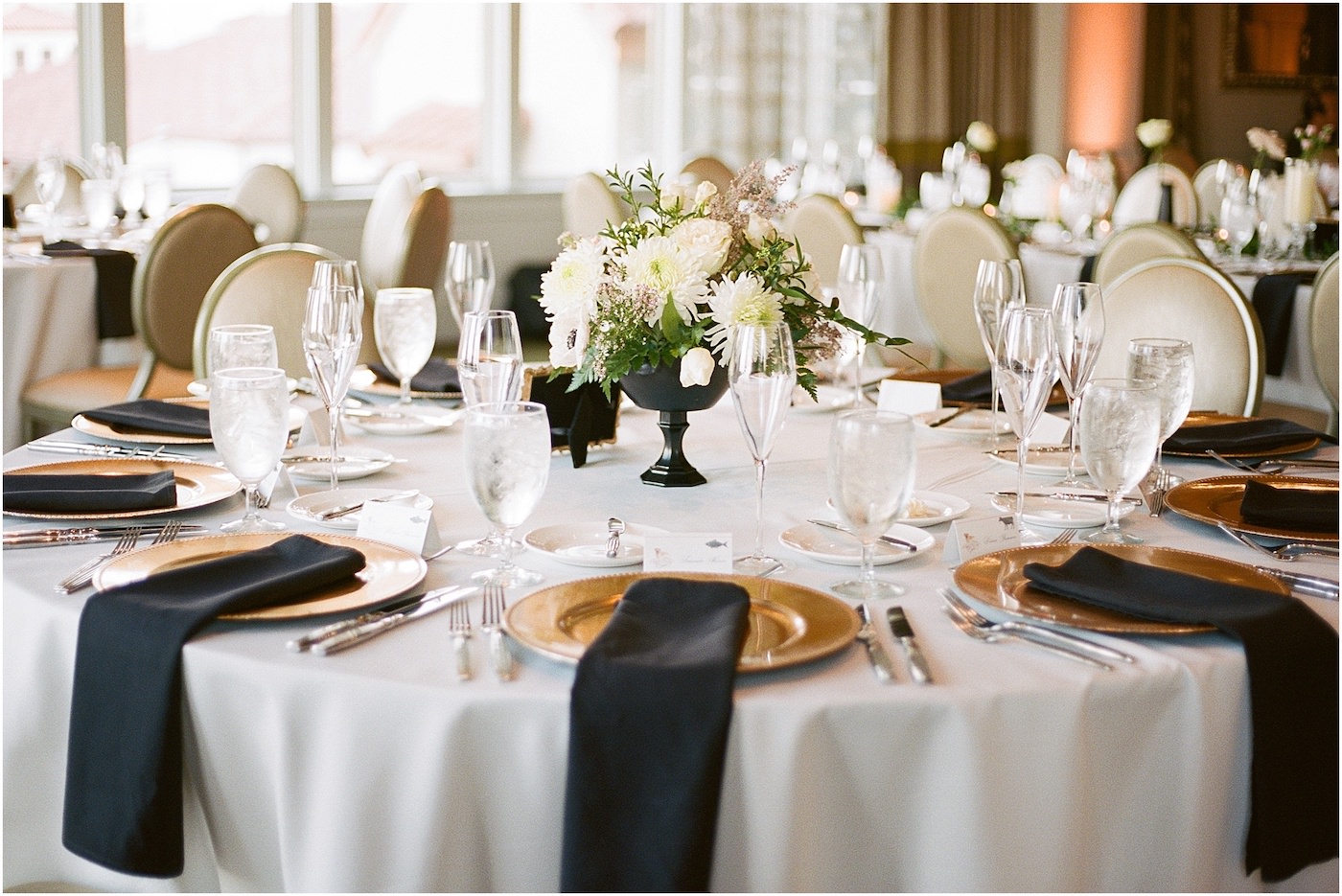St. Pete Wedding Venue The Birchwood | Indoor Ballroom Wedding Reception with White Table Linens and Black Napkins on Gold Charger Plates and White Floral Centerpieces with Greenery