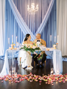 Romantic, Florida Bride and Groom at Wedding Reception with Decor, Ghost Acrylic Sweetheart Table with Light Blue Linens, Tall Taper Candles with Gold Accents, Vibrant Florals with Petals Scattered on the Floor, Whimsical Bridal Bouquet with Pink, Yellow, Peach and Green Florals, Blue Draping | Tampa Bay Wedding Planner Kelly Kennedy Weddings and Events | Kate Ryan Event Rentals | Tampa Wedding Venue The Orlo House