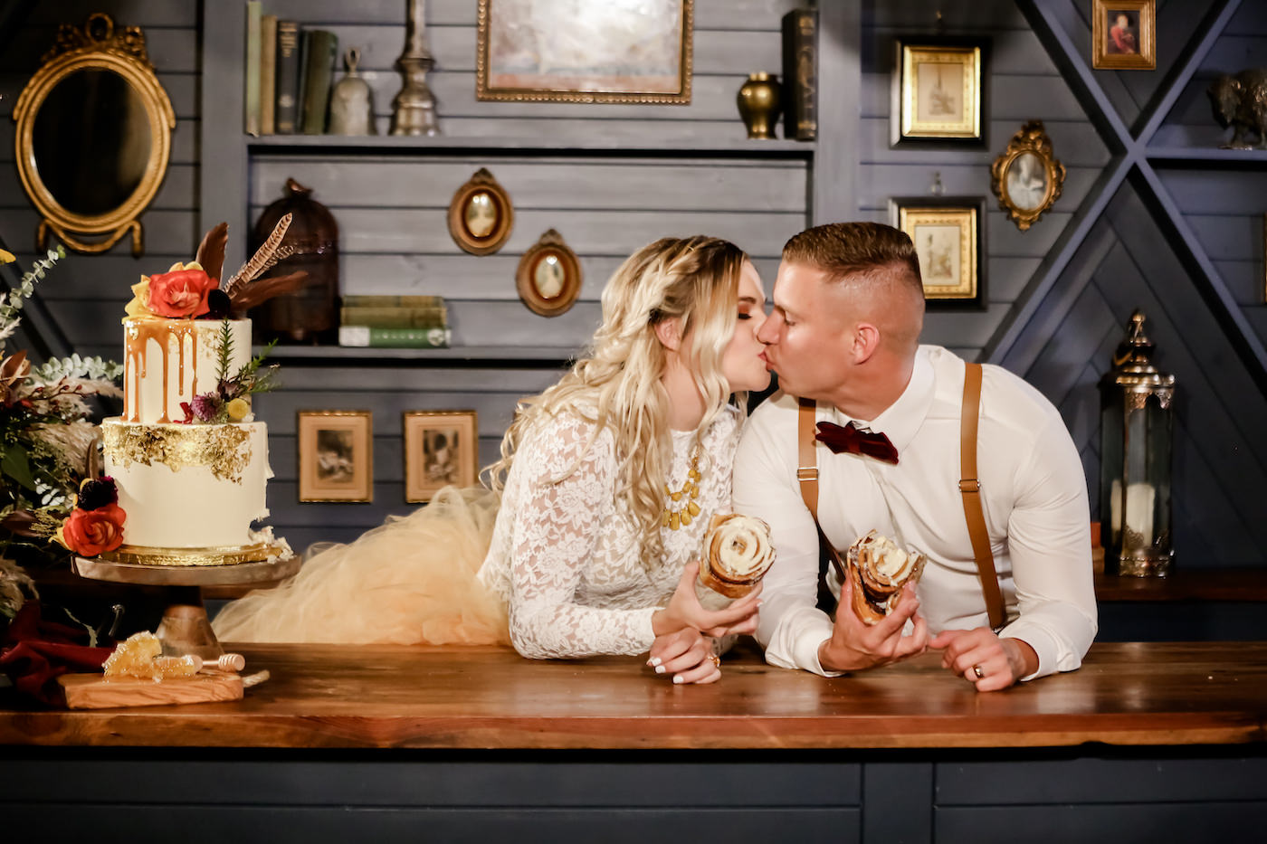 Florida Bride and Groom Kiss During Cake Cutting, Eating Bear Claws, Boho Inspired Bride Wearing Lace Long Sleeve Shirt with Blush Orange Tulle Skirt, Groom in Suspenders with Velvet Bowtie, Two Tier Bohemian Themed White Buttercream Wedding Cake with Honey Drip Icing, Edible Gold Glitter Detailing, with Autumn Florals, Deep Red Roses, Purple Thistle, Eggplant Florals, White and Gold Flowers, Greenery and Pampas Grass | Florida Wedding Cake Artist The Artistic Whisk | Downtown St. Petersburg Wedding Planner Blue Skies Weddings and Events | Florida Wedding Photographer Lifelong Photography Studio | Tampa Bay Unique Wedding Venue Station House in DTSP