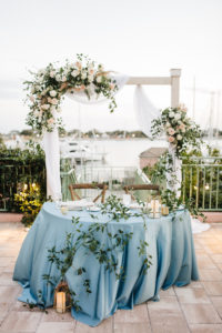 Bohemian Beach Inspired Wedding Reception and Decor, Sweetheart Tables with Soft Dusty Blue Table Linens, French Country Cross Back Wooden Chairs, Modern Geometric Candle Holders, In front of Ceremony Arch with Ivory Flowers and Blush Pink Roses, Pampas Grass Bouquets with Greenery, White Draping | Waterfront Wedding Venue The Esplanade Location of the Vinoy Renaissance Resort in Downtown St. Petersburg | Tampa Bay Luxury Wedding Planner Parties A’ La Carte | Rentals A Chair Affair And Over the Top Rental Linens | Wedding DJ Grant Hemond and Associates