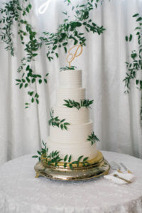Elegant Wedding Cake and Table at Wedding Reception, Love You More Neon White Sign, Four Tier White Buttercream Wedding Cake with Textured Frosting, Greenery Floral Accents, Gold Cake Stand, Last Name Initial Topper, Lace Linen Cake Tablecloth | Florida Wedding Planner Parties A'La Carte | Downtown St. Petersburg Wedding Florist Bruce Wayne Florals