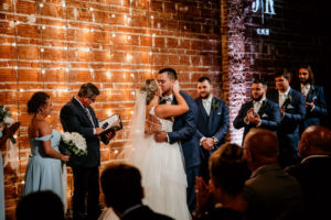 Downtown St. Pete Bride and Groom Intimate First Kiss in Indoor Summer Wedding Ceremony, Exposed Red Brick Wall with String Lighting | Tampa Bay Unique Wedding Venue NOVA 535 | Florida Wedding Photographer Bonnie Newman Creative