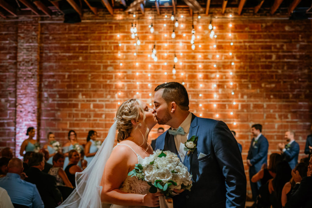 Downtown St. Pete Bride and Groom Intimate Kiss in Indoor Summer Wedding Ceremony, Exposed Red Brick Wall with String Lighting | Tampa Bay Unique Wedding Venue NOVA 535 | Florida Wedding Photographer Bonnie Newman Creative | Wedding Hair and Makeup Artist Femme Akoi Beauty Studio