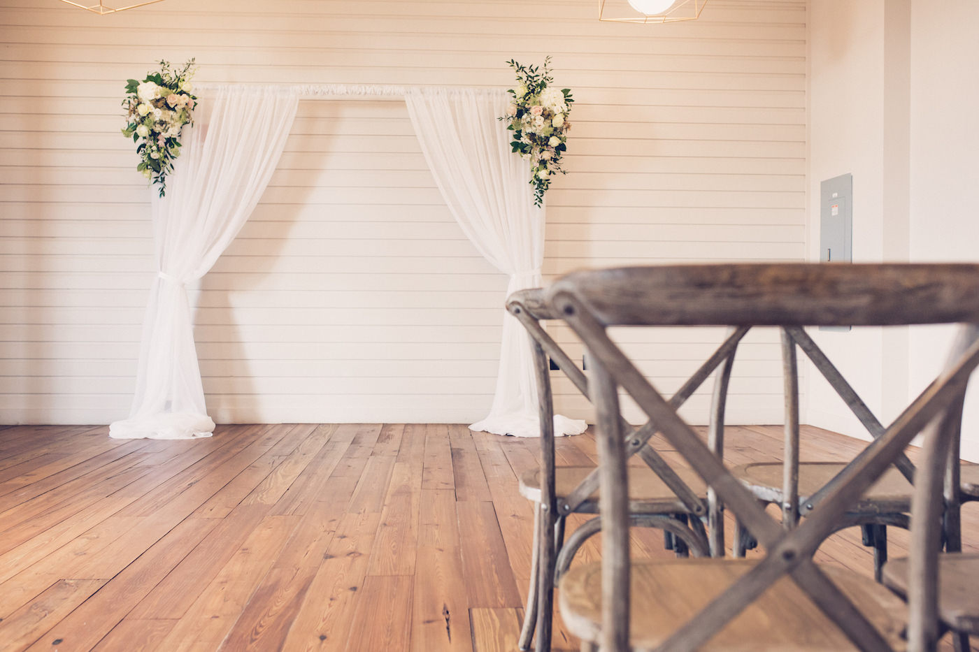 Indoor Tampa Wedding Ceremony with Pipe and Drape Fabric Arch Backdrop with Greenery and White Flower Arrangements and French Country Cross Back Wood Chairs