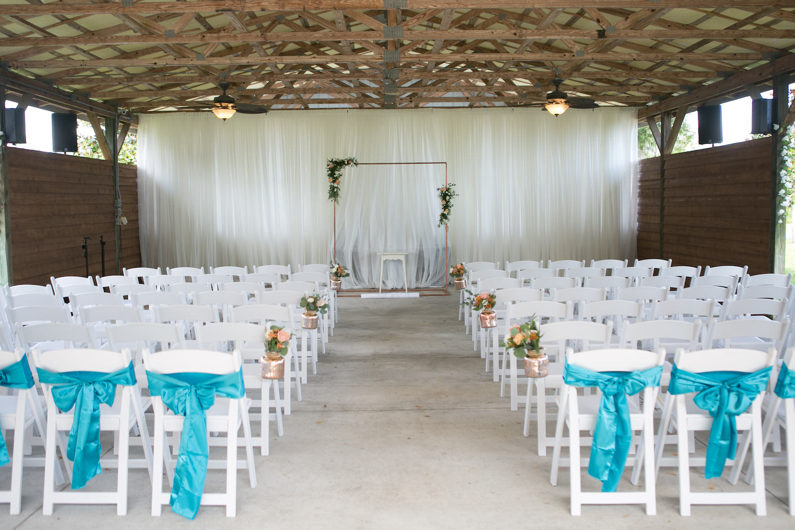 Simple Wedding Ceremony Decor, White Folding Chairs with Teal Blue Bows, Gold Mercury Jars with Floral Arrangements, Rectangular Copper Arch with Greenery Arrangements | Wedding Photographer Carrie Wildes Photography | Pinellas Park Wedding Venue Shahnasarian Hall | Wedding Linen Rentals Gabro Event Services