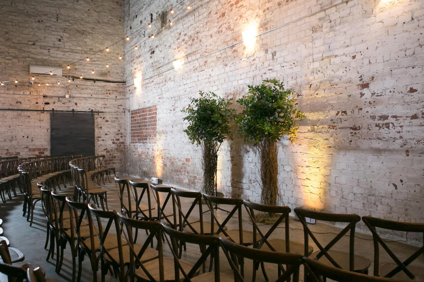 Wes Anderson Themed Wedding Ceremony Decor, Wooden Cross back Chairs, Curly Willow Trees and White Brick Wall with Hanging Lights | Wedding Photographer Carrie Wildes Photography | Tampa Bay Wedding Planner UNIQUE Weddings + Events | Wedding Florist Monarch Events and Design | Wedding Venue Rialto Theatre