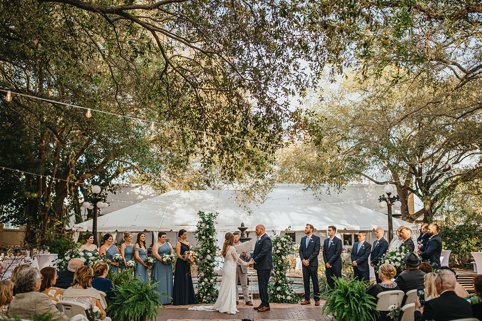 Bride and Groom Exchanging Vows during Ybor City Tampa Outdoor Historic Museum Garden Wedding Ceremony with Fountain Backdrop | Wedding Floral Arrangements with Classic White Roses and Lisianthus and Natural Greenery by Tampa Wedding Florist Monarch Weddings and Events | Dusty Blue Steel Grey Bridesmaid Dresses and Navy Blue Groomsmen Suits