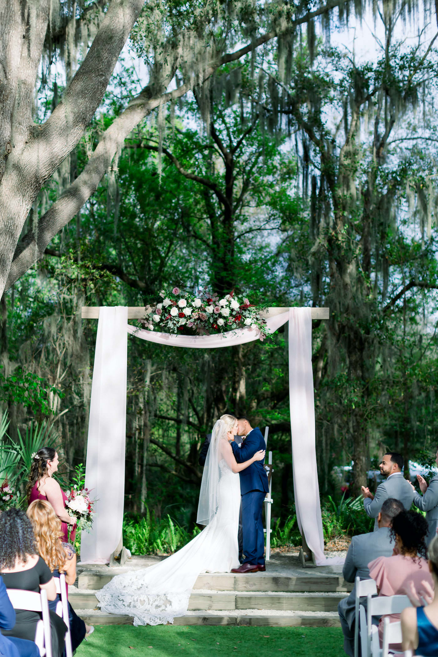 Bride and Groom First Kiss | Outdoor Florida Wedding Ceremony with Draped Arch and Burgundy and White Floral Spray Arrangement