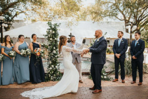 Bride and Groom Exchanging Vows during Ybor City Tampa Outdoor Historic Museum Garden Wedding Ceremony with Fountain Backdrop | Wedding Floral Arrangements with Classic White Roses and Lisianthus and Natural Greenery by Tampa Wedding Florist Monarch Events and Designs | Dusty Blue Steel Grey Bridesmaid Dresses and Navy Blue Groomsmen Suits