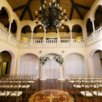 Tampa Wedding Venue Avila Golf & Country Club Indoor Ceremony with Gold Chiavari Chairs, Pipe and Drape Backdrop with Blush Pink and Ivory Rose Floral Arrangements with Greenery and Crystal Chandelier