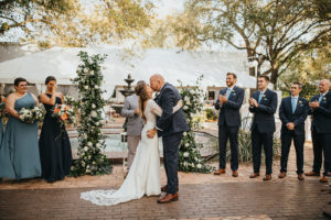 Bride and Groom First Kiss during Ybor City Tampa Outdoor Historic Museum Garden Wedding Ceremony with Fountain Backdrop | Wedding Floral Arrangements with Classic White Roses and Lisianthus and Natural Greenery by Tampa Wedding Florist Monarch Weddings and Events | Dusty Blue Steel Grey Bridesmaid Dresses and Navy Blue Groomsmen Suits