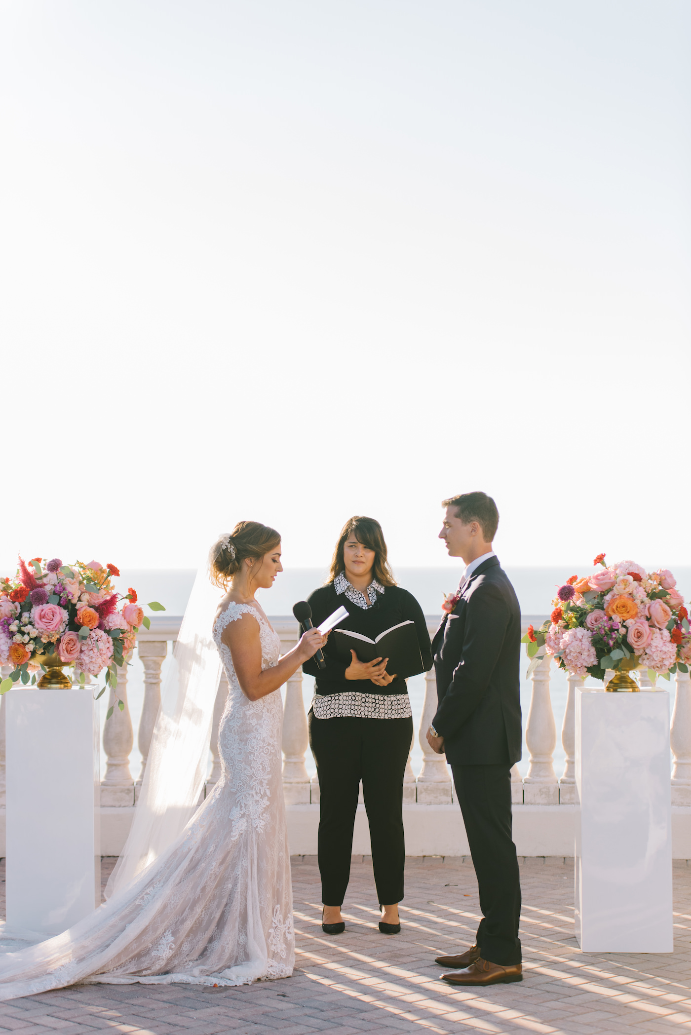 Bride and Groom Exchanging Vows during Outdoor Rooftop Ceremony at Clearwater Wedding Venue Hyatt Regency Clearwater Beach | Ceremony Backdrop White Pillars topped with Colorful Vibrant Tropical Floral Arrangements | Tampa Bay Wedding Photographer Kera Photography