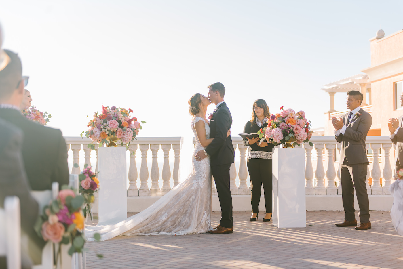 Bride and Groom First Kiss during Outdoor Rooftop Ceremony at Clearwater Wedding Venue Hyatt Regency Clearwater Beach | Ceremony Backdrop White Pillars topped with Colorful Vibrant Tropical Floral Arrangements | Tampa Bay Wedding Photographer Kera Photography