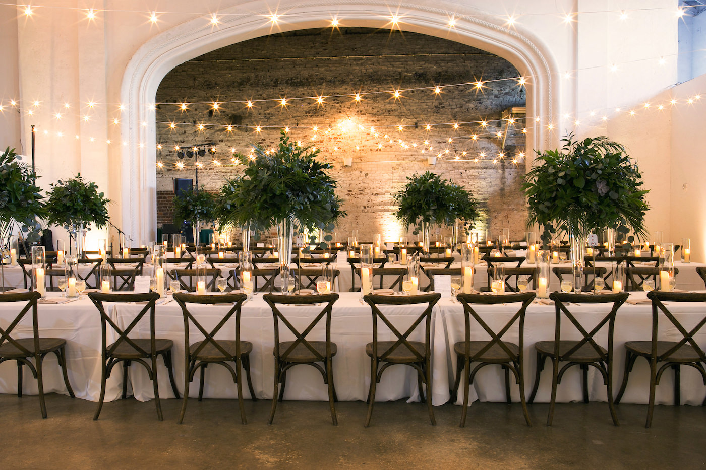 Wes Anderson Inspired Wedding Reception Decor, Long Feasting Tables with Wooden Cross back Chairs, Tall Greenery Plants Centerpieces and String Lights, Hurricane Glass Candles | Wedding Photographer Carrie Wildes Photography | Tampa Bay Wedding Venue The Rialto Theatre | Wedding Planner UNIQUE Weddings and Events | Wedding Florist Monarch Events and Design | Olympia Catering