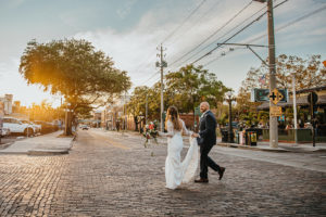 Ybor City Tampa Wedding Bride and Groom Outdoor Cobblestone Street Sunset Portrait | Long Sleeve Lace Bridal Gown Wedding Dress | Classic Navy Blue Groom Suit | Organic Natural Loose Wedding Bridal Bouquet with Coral Pink Peonies and White Roses with Succulents and Eucalyptus Greenery by Tampa Florida Monarch Events and Designs
