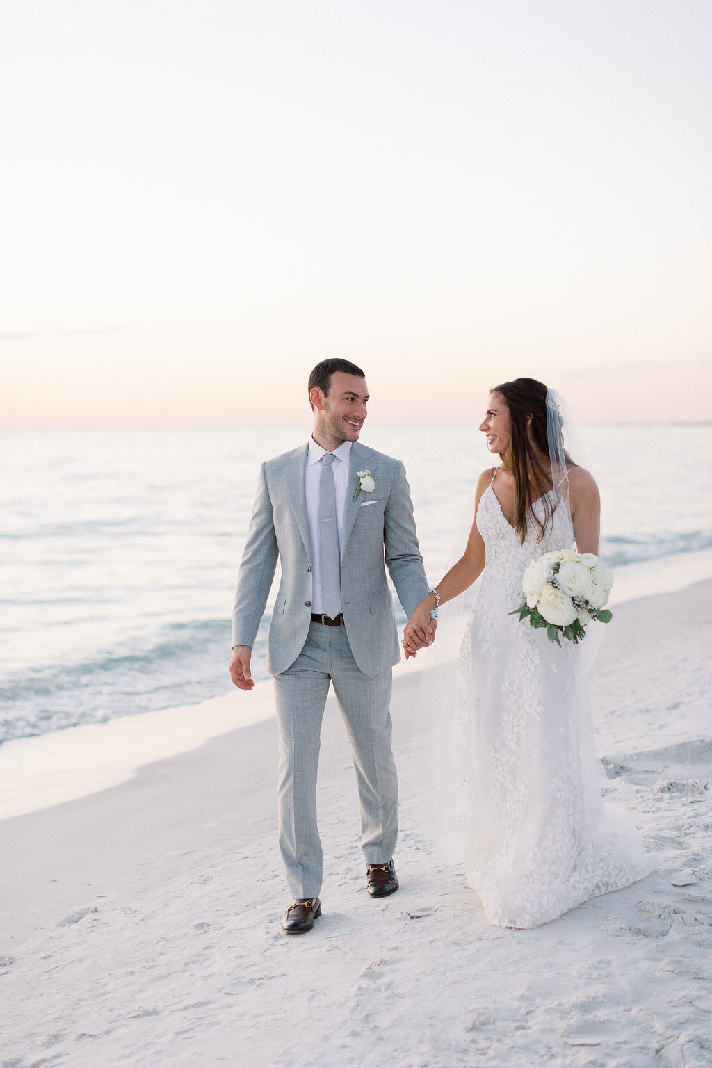 Classic Florida Bride and Groom Hold Hands and Walk on the Sand on Gulf Coast Beach, Bride Wearing Lazaro Wedding Dress Holding Ivory and White Floral Bouquet, Groom in Silver Suit, Sunset Wedding portrait