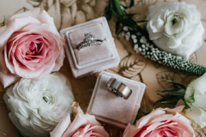 Wedding Rings Shot with Diamond Solitaire Engagement Ring and Platinum Mens Band in Blush Pink Velvet Boxes with Fresh Roses and Ranunculus