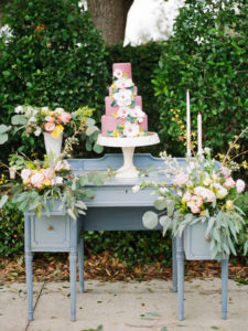 Vintage Furniture Desk Wedding Cake Table with Pastel Pink and Yellow Flowers and Greenery | Four Tier Colorful Spring Wedding Cake with Pink Fondant Icing and Sugar Flower Magnolias on a Round Pedestal Stand